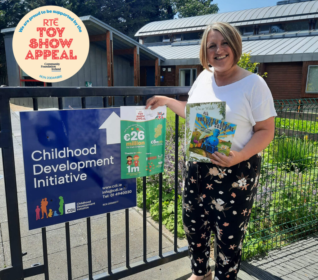 Denise Lake, Literacy and STEAM Co-ordinator, standing at the gate at CDI, in the sunshine, holding children's books and a poster for the RTE Toyshow